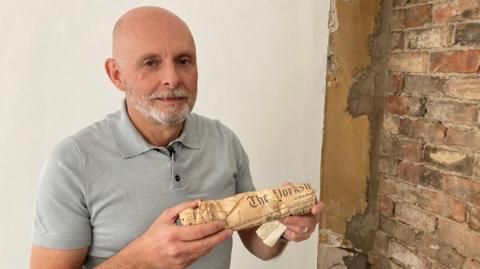 A man is smiling at the camera. He is wearing a grey top and has a grey beard. He's holding up a package which is covered in a stained newspaper. He's standing next to a brick wall.