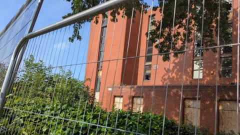 A red brick building behind fencing which has boarded up and smashed windows