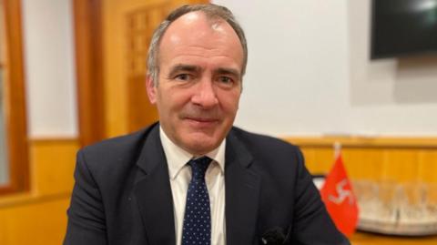 Chief Minister Alfred Cannan is wearing a black suit and dark spotted tie, in his wood panelled office, with a small Manx flag to his right.