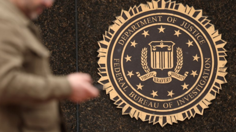 A man whose face is blurred walks past an FBI crest that is on the side of a building. 