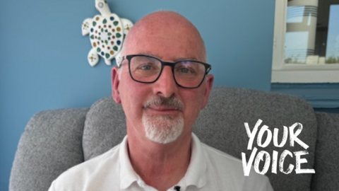 A bald man with black glasses and a white polo shirt. He is sat in a grey chair in front of a light blue wall and looking into the camera.