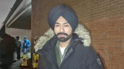 Tarsem Singh Jassar wearing a blue turban and blue coat. He has a black beard. He is wearing a white t-shirt, grey shirt and grey top. There is a brick wall underneath a bridge behind him. There are two yellow traffic cones and members of the cast and crew stand in the background.