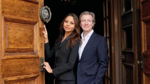 The 8th Marquess and Marchioness of Bath stand together at the large wooden doorway to Longleat House. They are smartly dressed in dark suits and are smiling at the camera