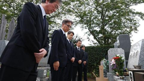 Five men wearing dark suits bow before a grave in Japan. The grave is made of grey marble, and has flowers in a vase in front of it