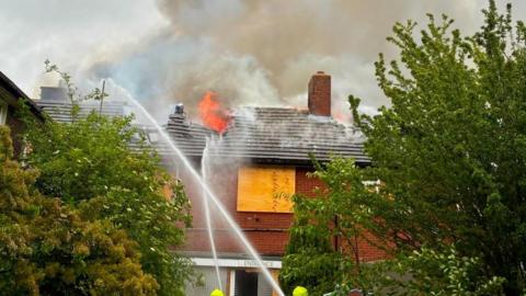 Water jets being sprayed onto a large red brick building, with trees either side of it. Its windows are boarded up and there is a sign above the front door. Flames are coming out the roof and parts of the roof have collapsed. 