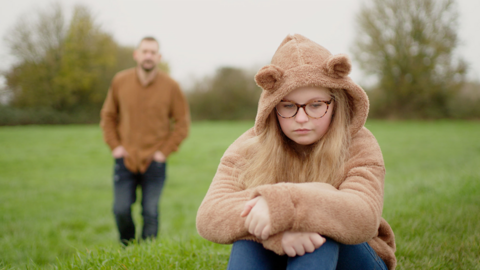 A thoughtful-looking girl in the foreground wearing a brown hoodie with bear ears, with an out-of-focus man in the distance walking towards her