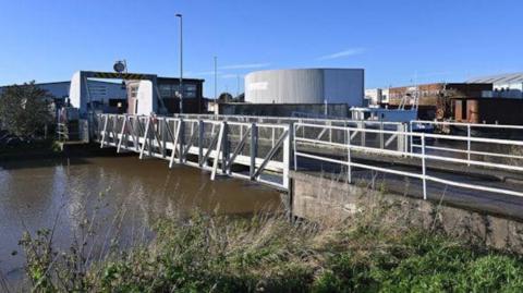 A bascule bridge over murky water. Industrial buildings can be seen in the distance. The sky is blue.
