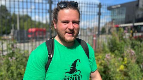 A bearded man with a green t-shirt and sunglasses on his head, standing in front of green bushes and railings