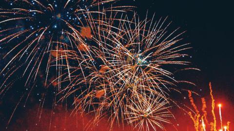 Starbursts of orange and yellow fireworks against a dark sky, with red smoke and lights rising to the right.