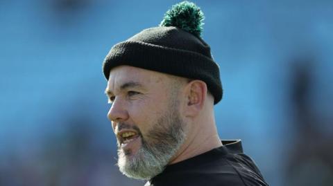 Bearded Northampton Saints assistant coach Matt Ferguson looks on during a game, while wearing a bobble hat.