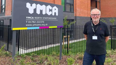 Daniel Flynn, a man with receding white hair and wearing a black polo shirt and dark blue jeans, is stood outside a block with windows and different floors, next to a grey sign which has white lettering that reads YMCA North Staffordshire.