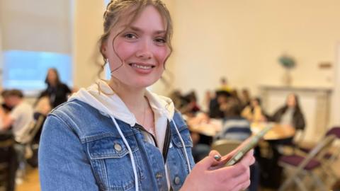 A teenage girl stands holding a phone and smiling at the camera. There are other students sitting at tables, blurred in the background.