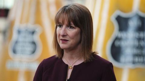 Rachel Reeves stood in front of a yellow backdrop with black logos on it. 