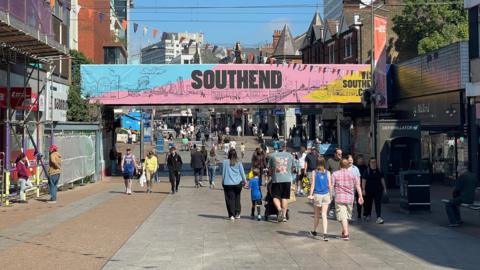 People walking up and down the pedestrianised Southend High Street. There is a large overhead banner saying SOUTHEND on a blue, pink and yellow background