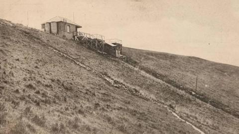 An old postcard in sepia. It shows a lonely building on top of a hill. 