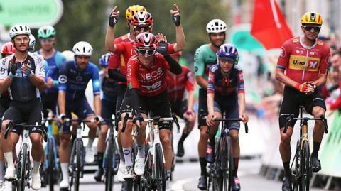 Geraint Thomas crosses the finish line cheered by the riders in his farewell and last race as a professional cyclist during the 21st Tour of Britain 2025. The riders are all wearing cycling colours and helmets.