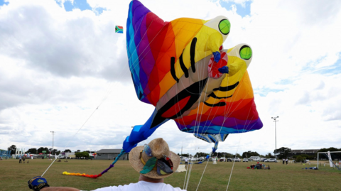 Woman wearing straw hat flies a kite that looks like an animal in rainbow colours 