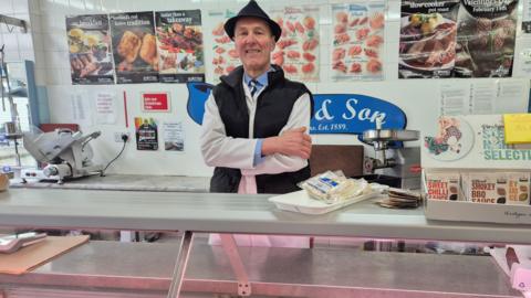 James Baillie stands behind the counter of a butcher's shop. He is wearing his work clothes, while posters behind him advertise the stop's meats and products. 