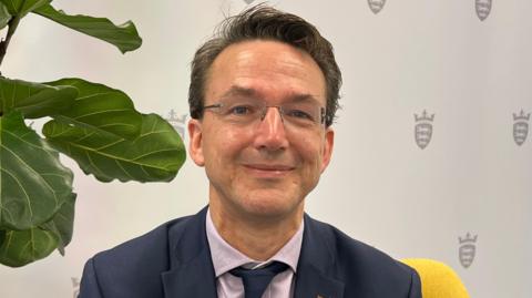 A man wearing a suit and tie sitting on a yellow chair with a plant to his left and a white background with the States of Jersey emblem behind.