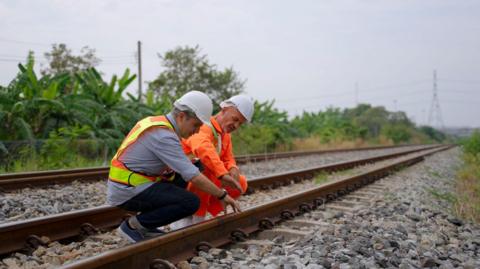 The image shows two engineers working on the rail tracks. They are bending down an inspecting a track.