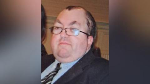 Graham Milliken looking straight at the camera. He has short, receding dark hair and glasses. He is wearing a suit jacket with a blue shirt and a striped tie