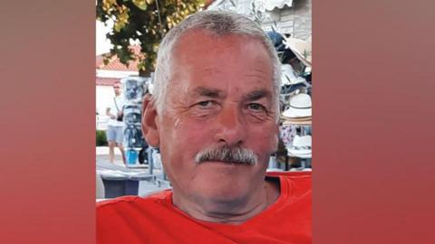 A man with short grey hair and a grey moustache and wearing a red T-shirt is smiling while sitting in the sun. Behind him is what appears to be an outdoor shop display of sun hats as well as a tree.