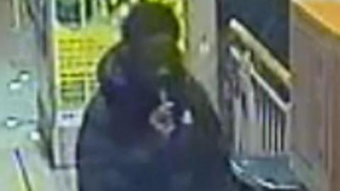 CCTV of a man dressed in a black coat holding something in his hand which is in his mouth near a turnstile at a Metrolink tram stop.