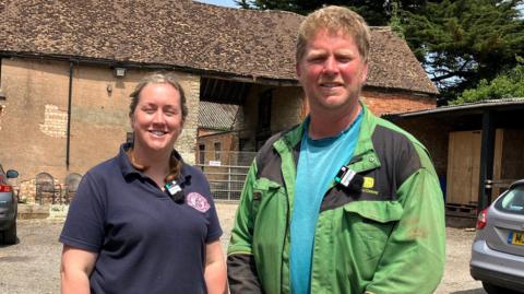 Heather Clarke (left) stands next to her husband George. She is wearing a blue top and plaits in her hair. Geoorge wears a lighter blue top and green and black coat. 