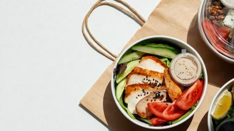 A takeaway salad in a paper bowl on a brown paper bag with wooden fork