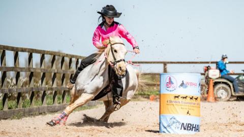 A girl wearing a cowboy hat, pink shirt and black trousers rides a grey horse at speed around and blue and yellow barrel, in a sandy arena surrounded by a fence