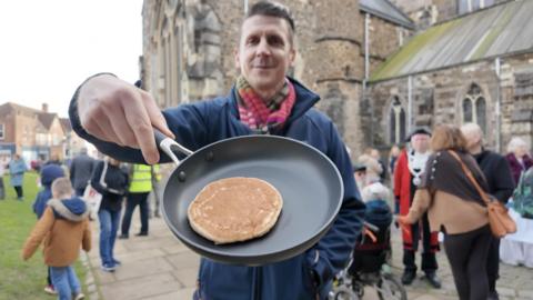 A man smiling holding a frying pan with a small pancake.