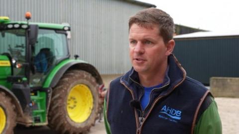 Ashley Jones wears a gilet over a green top and stands side on to the camera talking to an out of shot journalist with a serious expression. Behind him to the left is a large green tractor with yellow wheels.