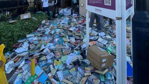 Piles of books cover the pavement outside Broad Green Library in Croydon, many in good condition and some in open boxes. Two people are visible among the stacks, with a black skip to the left and a library sign in the foreground.
