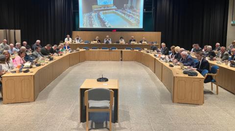A large meeting room with participants seated around a U‑shaped table. A single chair with a microphone sits in the centre. At the front, a panel faces the room, and a large screen above them displays a live view of the meeting. The room is well‑lit with wood‑panel desks and multiple people using laptops and documents.