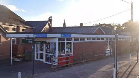 A single-storey building with a wooden fence on one side and a porch and a road along the right.