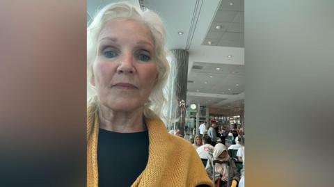 Lenise Blom, who is wearing a yellow jumper and black shirt takes a selfie while stood inside the terminal at Dubai Airport. Dozens of people are sat behind her. She is a woman in her 70s who has wavy white hair.