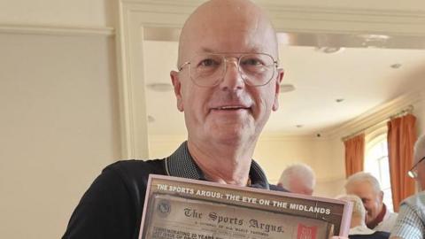 Norman Bartlam is wearing glasses and a dark shirt. He is holding his book up which is call The Sports Argus: The Eye on the Midlands