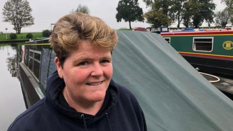 Tracey Baxter MD at Fox Narrowboats has red short permed hair with a dark blue hoodie, behind her are narrowboats with trees visible on the river bank