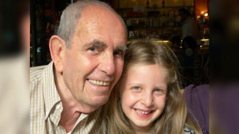 Dr George Garai is on the left, and Ella is on the right. Both are smiling at the camera, and appear to be in a cafe or restaurant. Dr Garai is elderly with short grey hair, and Ella is six or seven years old with long blonde hair. 