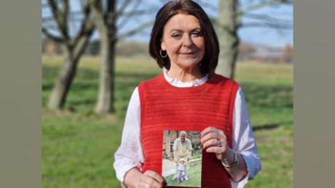 Carol Malia holding a photograph of her Dad Michael. She is wearing a red tank top and a white long-sleeved shirt and is standing in a sunny field with trees behind her.