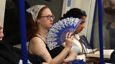 A woman with a bandana holding a blue fan sitting on the London underground