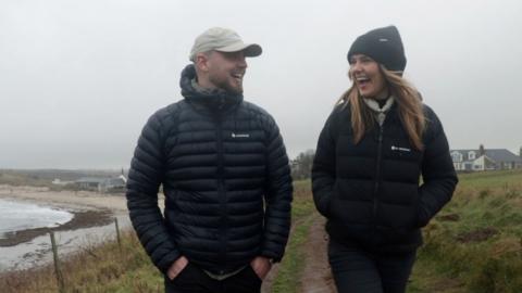 Scott Gillan and Steph Robinson, walking side by side smiling at each other, with a field and a beach in the background.