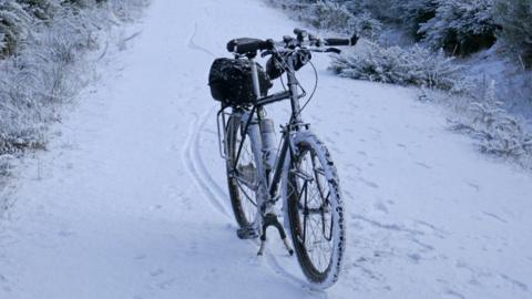 A snow covered bike without a rider on its stand on a snowy path