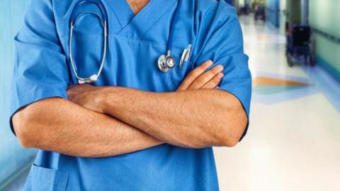 The torso of a nurse, wearing blue scrubs, with his arms crossed. He is standing in a hospital corridor. 