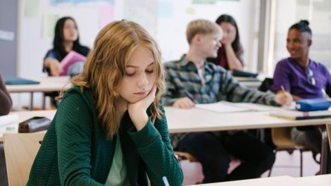 Young students in a classroom (stock photo). A young girl with shoulder-length fair hair is in the foreground wearing a green jacket and top.  She is looking down and resting her face on her chin.  Four other students are sitting in the background, two of whom are having a conversation.