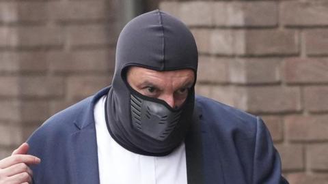 Richard Lester wearing a black balaclava to cover his face outside court. Only his eyes and part of his nose can be seen. He is wearing a navy suit jacket and white shirt in front of a brick building.