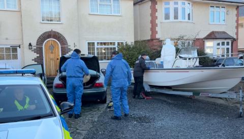 Police officers search the boot of a car and the open deck of a boat that are parked on a drive outside a house. An officer sits inside a police car in the foreground of the photo, the car being searched is in the middle of the frame, the boat is to the right. A neighbouring house can be seen on the other side of the boat