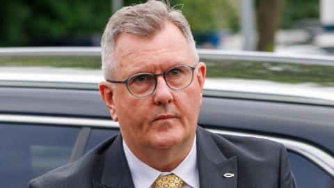 A man who has just come out of a car with a neutral expression on his face. He is wearing glasses and has short cropped, slightly thinning greyish hair. He is wearing a dark blue suit jacket, white shirt and yellow patterned tie. 