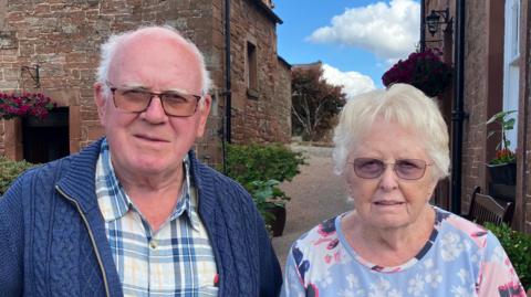 Chris and Ann Stevenson stand outside between two stone houses. They are both wearing glasses and have grey hair. Ann is wearing earrings. Chris wears a blue knitted cardigan over a blue-and-white checked shirt, while Ann wears a light blue floral top. Potted plants and hanging flower baskets can be seen by the houses and a cobbled pathway is seen between the buildings under a blue sky with clouds.