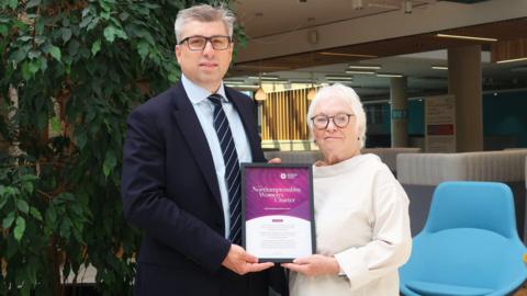 A man and a woman are holding a framed certificate. The man is wearing a suit and the woman is wearing a white top. There is a blue chair behind them and a large indoor plant 